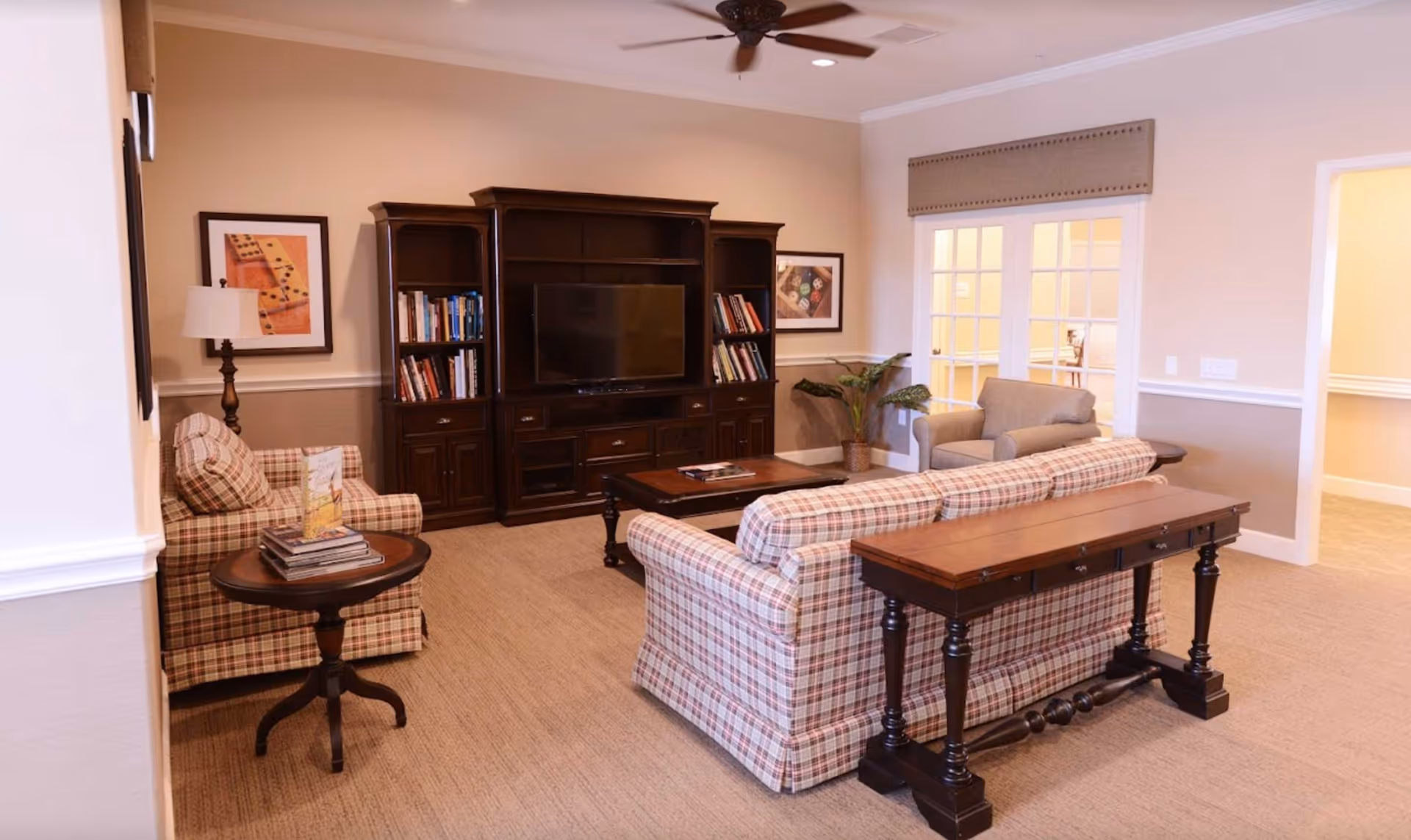 A cozy living room with plaid upholstered sofa and armchair, a wooden coffee table, a dark wood entertainment center with a TV and bookshelves, a side table with books and a lamp, beige walls with framed artwork, and a ceiling fan.