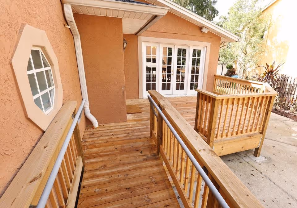 Wooden wheelchair accessible ramp leading to a set of white French doors on a peach-colored building exterior, with a small octagonal window on the left wall and some plants visible on the right side.