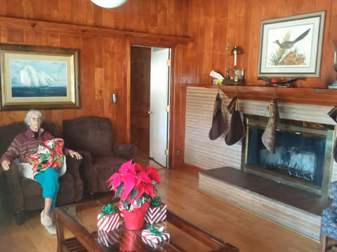 An elderly woman sitting on a brown armchair in a cozy living room with wood-paneled walls. There is a glass coffee table with a red poinsettia plant and wrapped gifts on it. The room features a brick fireplace with three stockings hanging and a framed bird picture above it. Another framed painting of a sailboat is on the wall behind the woman.