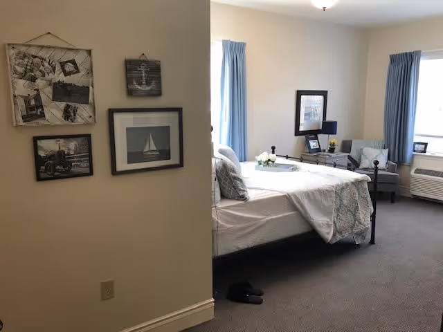 Neat furnished bedroom in a senior living facility featuring a made bed, armchair, wall art, and windows with blue curtains.