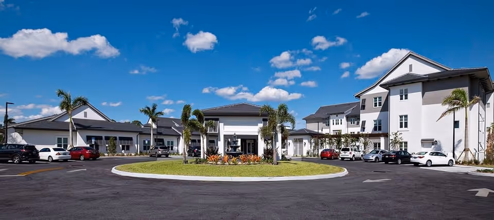 Exterior view of a senior living facility with multiple connected buildings, palm trees, a circular driveway with a landscaped center, and several parked cars under a blue sky with scattered clouds.