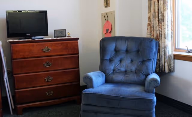 A cozy corner of a room featuring a blue upholstered armchair next to a wooden chest of drawers with a flat-screen TV and a digital clock on top. There is a window with patterned curtains allowing natural light into the room.