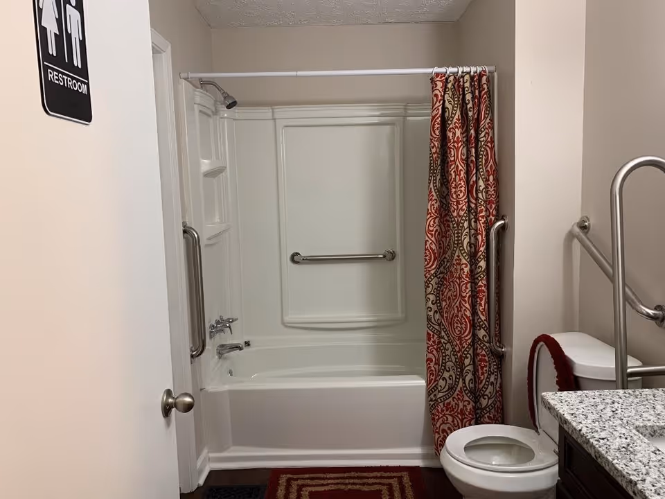 Interior view of a restroom with a white bathtub and shower combination. The shower has a silver grab bar and a showerhead. A red and beige patterned shower curtain is partially drawn to the side. There is a white toilet with a red cover on the tank and a silver grab bar next to it. A granite countertop with a sink is visible on the right side. The door to the restroom has a sign indicating it is a restroom for both men and women.