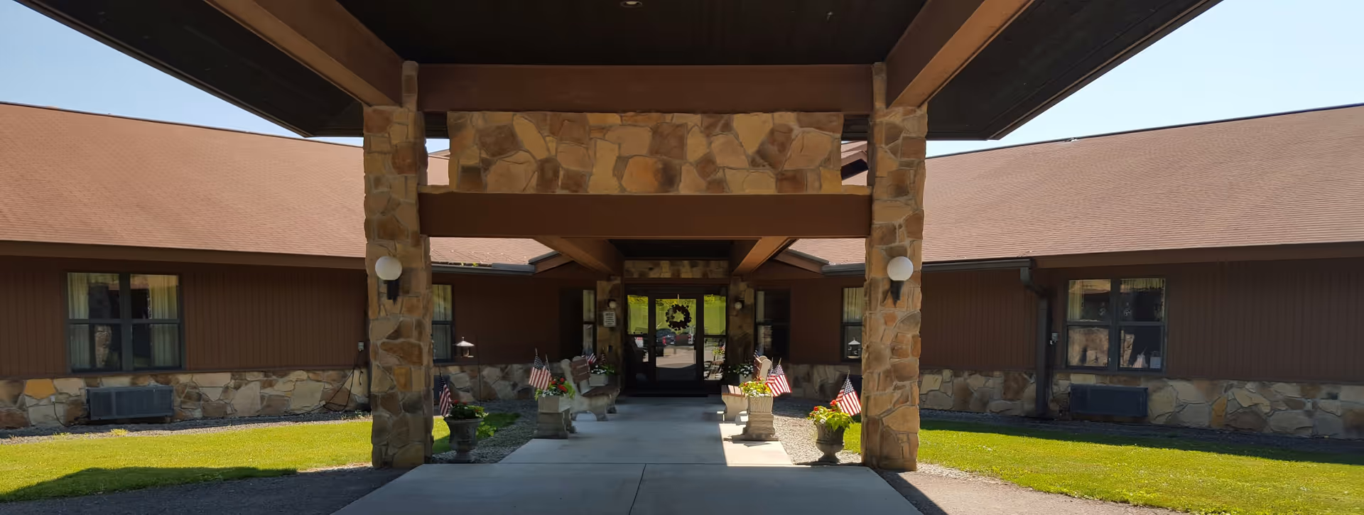 Covered stone-pillared entrance to a single-story senior care building with potted plants and small American flags lining the walkway.
