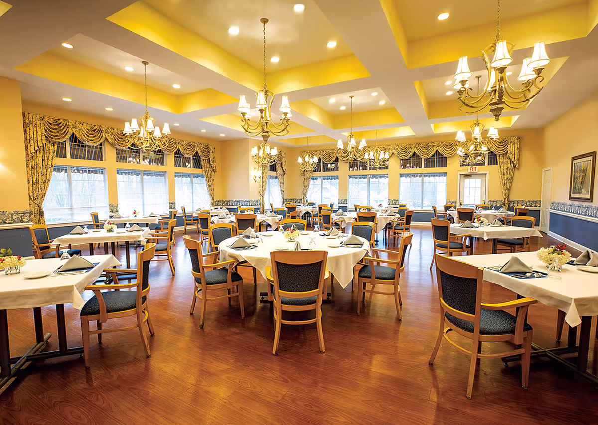 A spacious and well-lit dining room with multiple round and rectangular tables covered with white tablecloths, each set with napkins, plates, and floral centerpieces. The room features wooden chairs with blue cushions, large windows with patterned curtains, and elegant chandeliers hanging from a coffered ceiling. The floor is wooden, and the walls are painted in warm tones with decorative trim.
