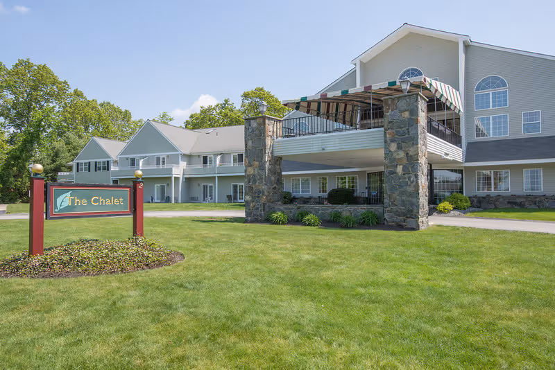 Exterior view of a senior living facility named The Chalet, featuring a well-maintained lawn, a two-story building with large windows, and a covered balcony supported by stone pillars under a clear blue sky.