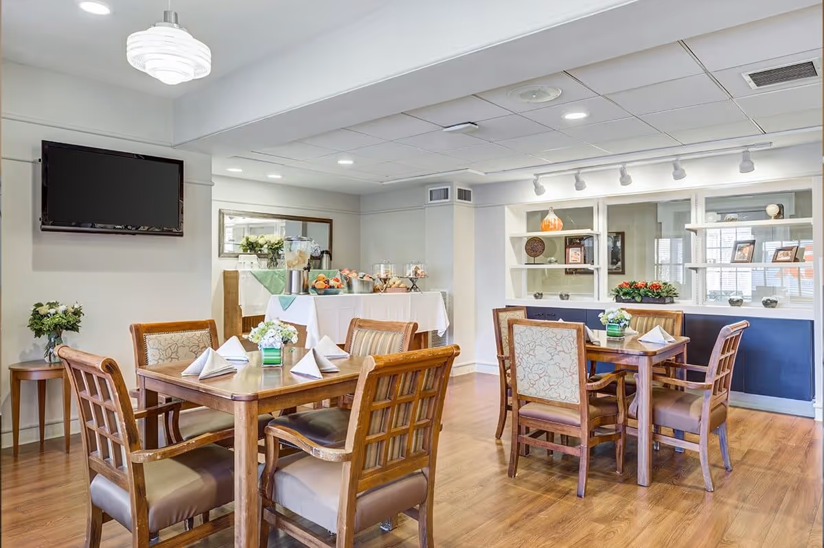A dining room with wooden tables and chairs, each table set with folded white napkins and small floral centerpieces. A buffet table with food and drinks is visible in the background, along with a wall-mounted flat screen TV and decorative shelves with framed pictures and ornaments. The room has wooden flooring and white walls with recessed lighting.