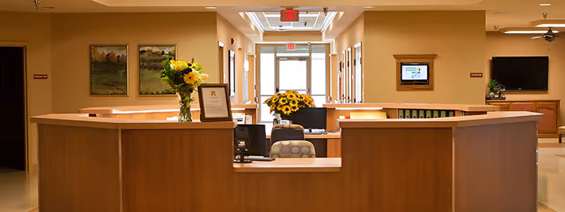 Wooden reception desk in a senior living lobby with sunflowers, seating, wall art, and a hallway leading to exit doors.
