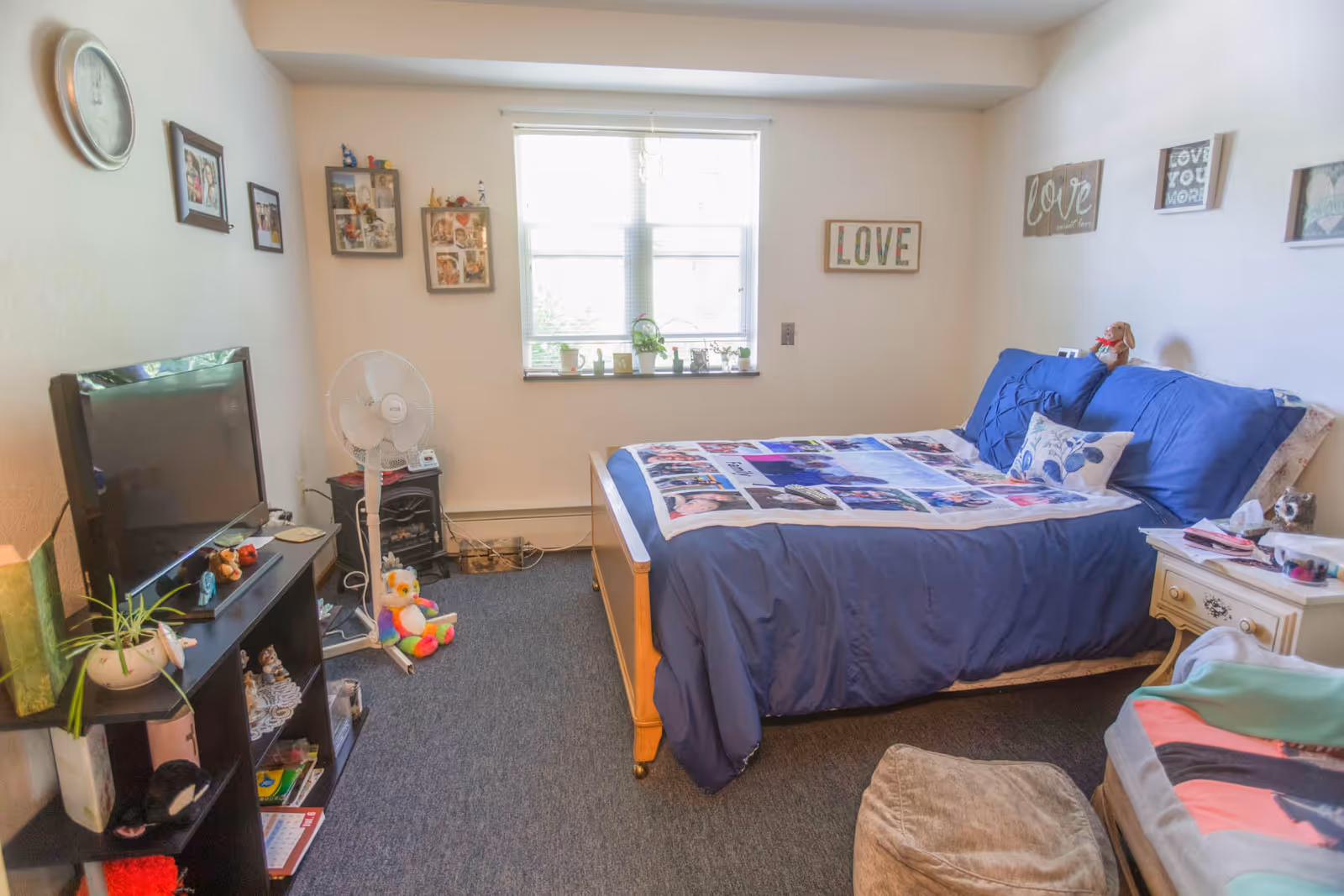 A cozy furnished bedroom with a bed dressed in blue bedding, a TV on a shelf, a window above the bed, and wall decorations.
