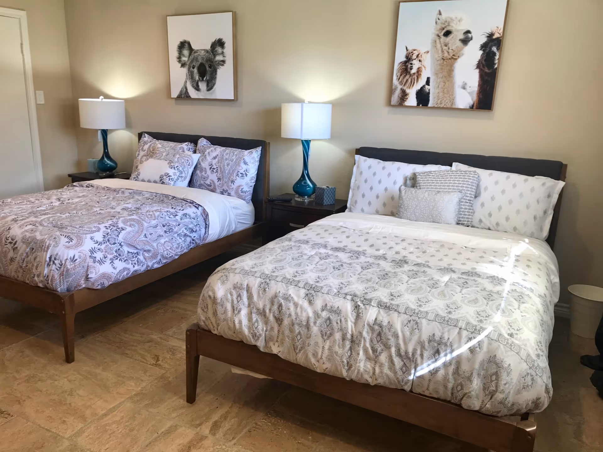 A bedroom with two beds, each with patterned bedding and multiple pillows. Between the beds is a nightstand with a blue lamp and a tissue box. Above each bed hangs a framed animal picture, one of a koala and the other of several llamas. The room has beige walls and a tiled floor.