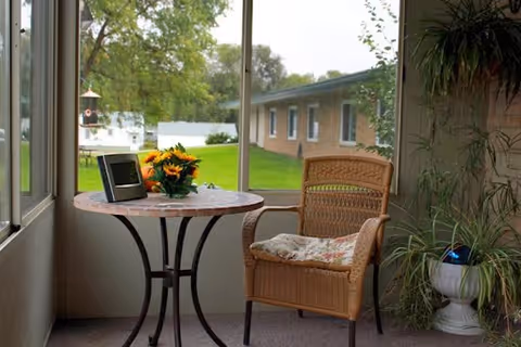 Sunroom seating area with a wicker chair and round table holding a bouquet and a tablet, looking out through large windows to a grassy yard and adjacent building.