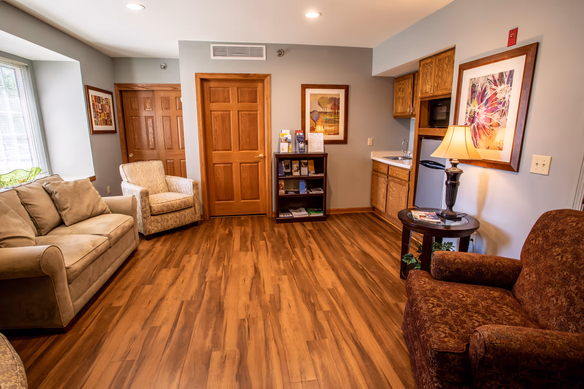 A bright senior living common room with sofas and armchairs, wood-look flooring, a small kitchenette, side table with lamp, and framed artwork.