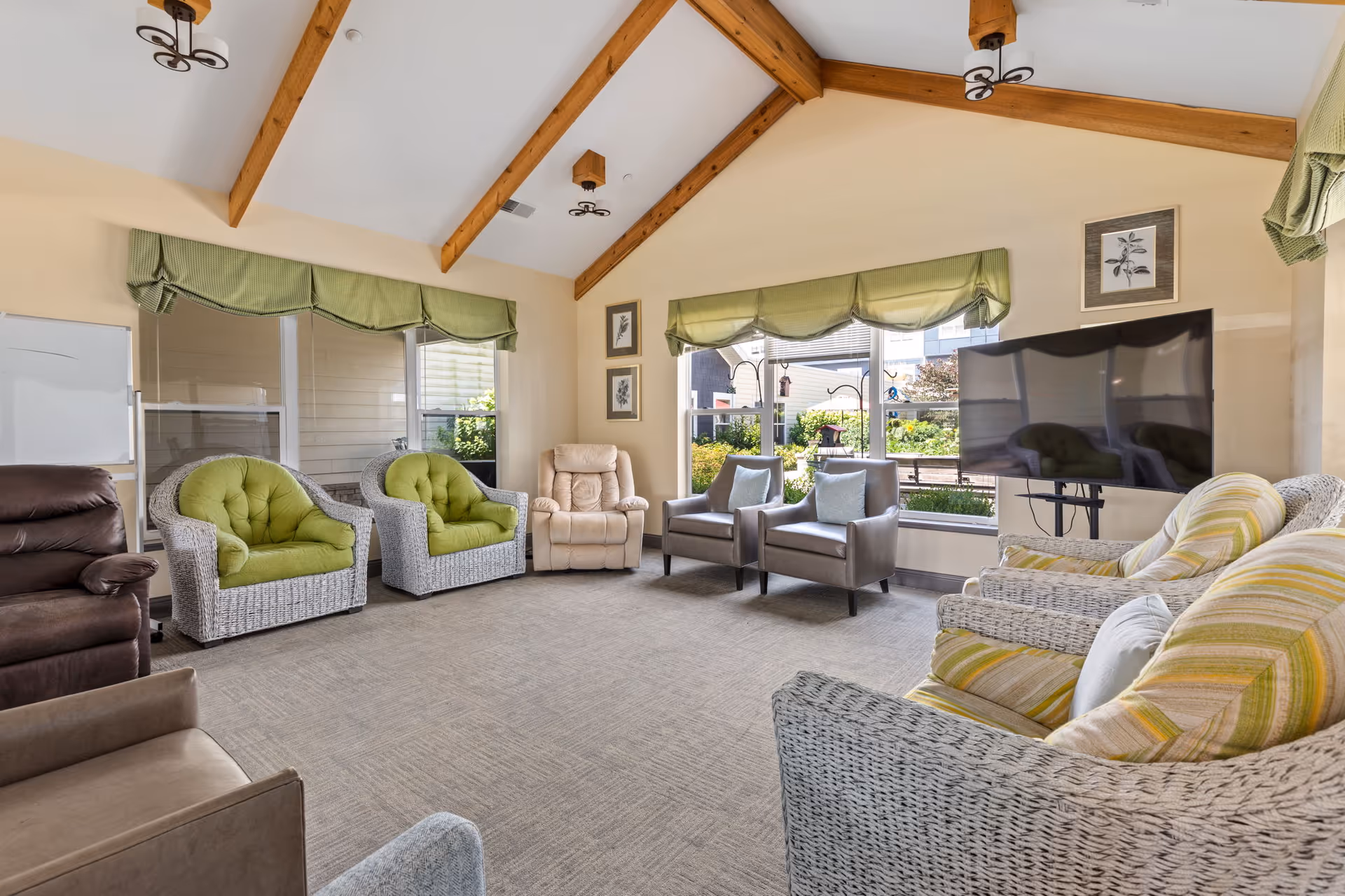 Bright communal living room with wicker and upholstered chairs arranged in a circle around a wall-mounted TV beneath a vaulted ceiling with wooden beams.