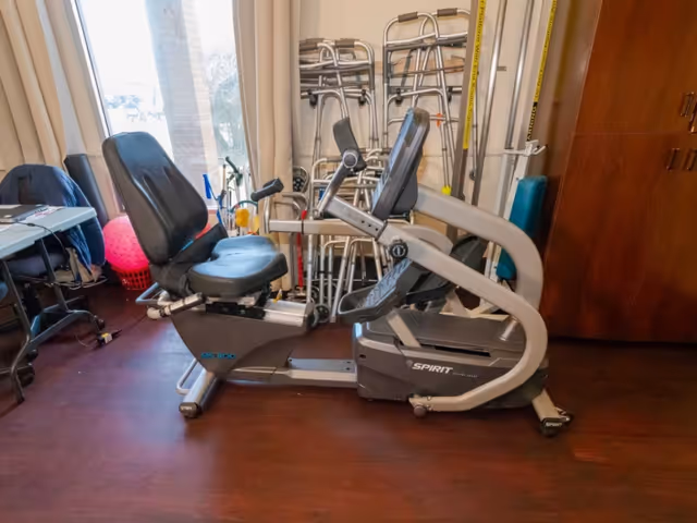 A recumbent exercise bike in a room with wooden flooring. Behind the bike, there are several walkers stacked against the wall. To the left, there is a table with a chair and some items on it, and a window with curtains letting in natural light. On the right side, there is a wooden cabinet.