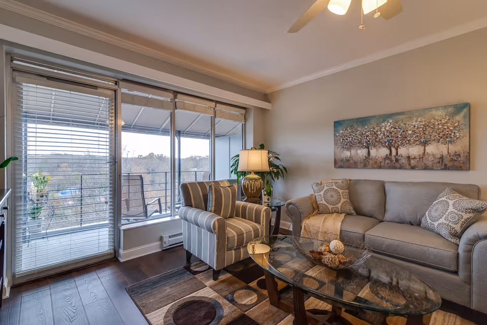 A cozy living room with a gray sofa adorned with patterned pillows and a throw blanket, a striped armchair, a glass coffee table with decorative items, a table lamp, and a large painting of trees on the wall. Large windows and a glass door with blinds lead to a balcony with outdoor chairs, overlooking a wooded area.