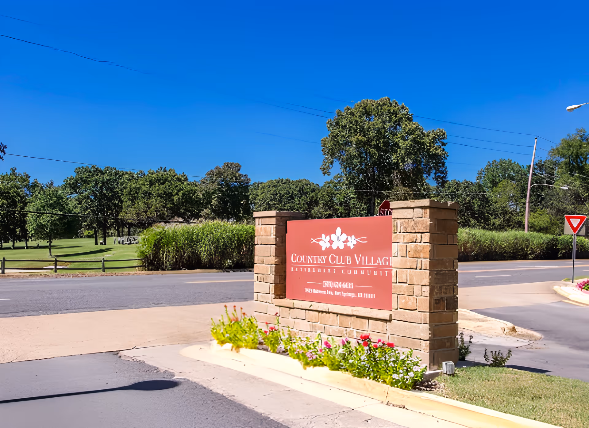 Entrance sign for Country Club Village Retirement Community made of brick with a red plaque displaying the community name, surrounded by flowers and greenery, with a clear blue sky and trees in the background.