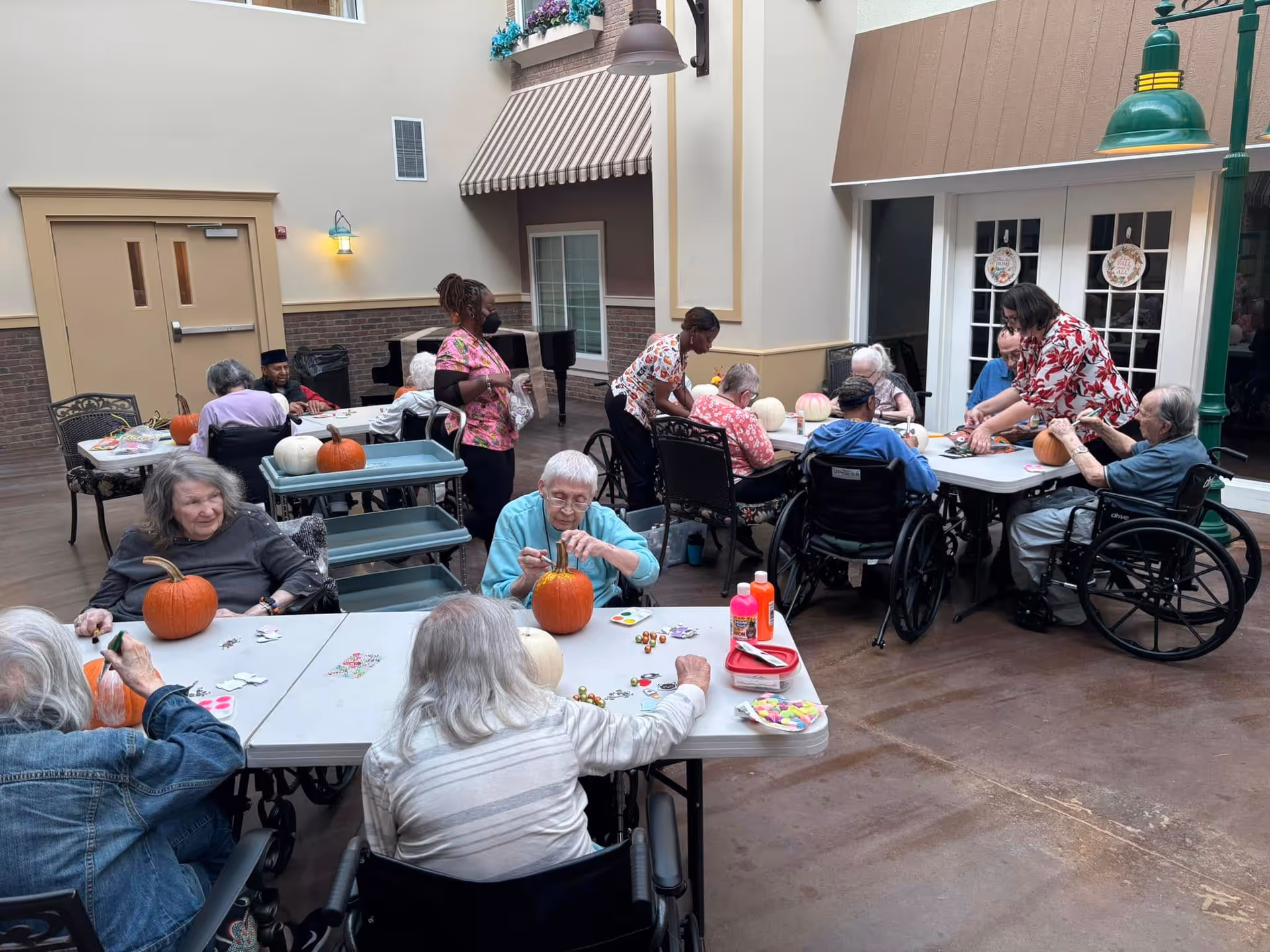 Elderly residents and staff sit around tables in an indoor common area decorating pumpkins.