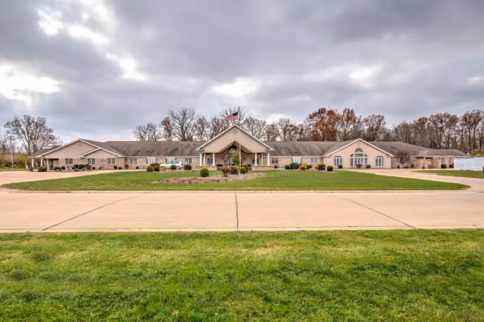Single-story senior living facility front with a flagpole, landscaped lawn and circular driveway under a cloudy sky.