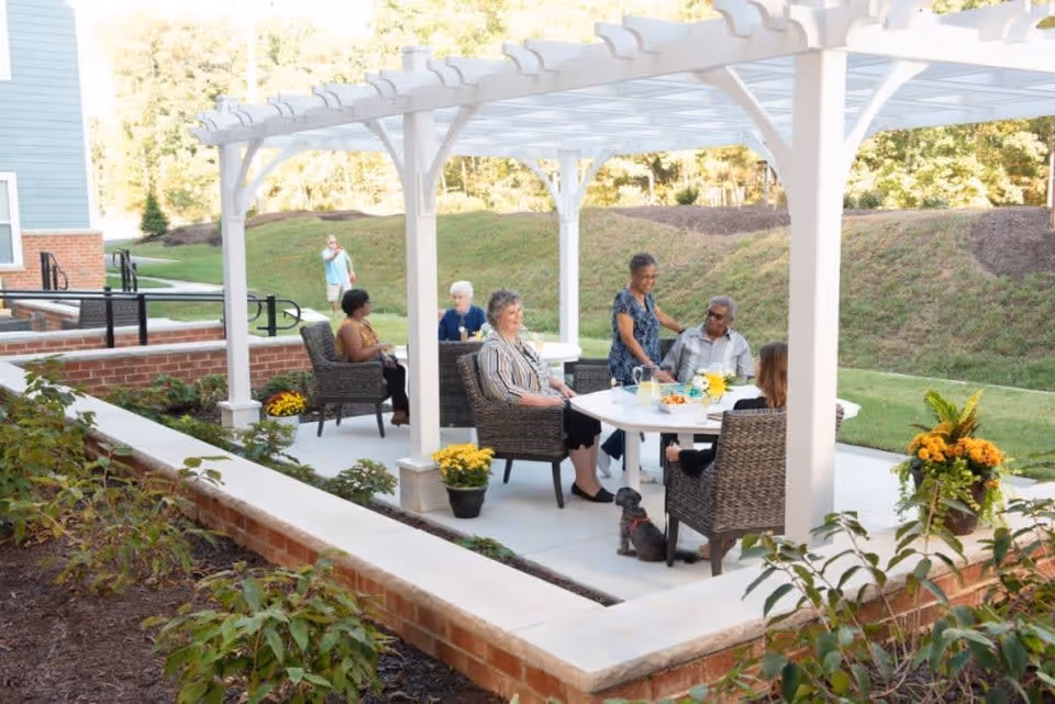 A group of elderly people sitting and socializing under a white pergola in an outdoor patio area with wicker chairs and a white table. There are potted yellow flowers and greenery around the patio, with a grassy hill in the background.