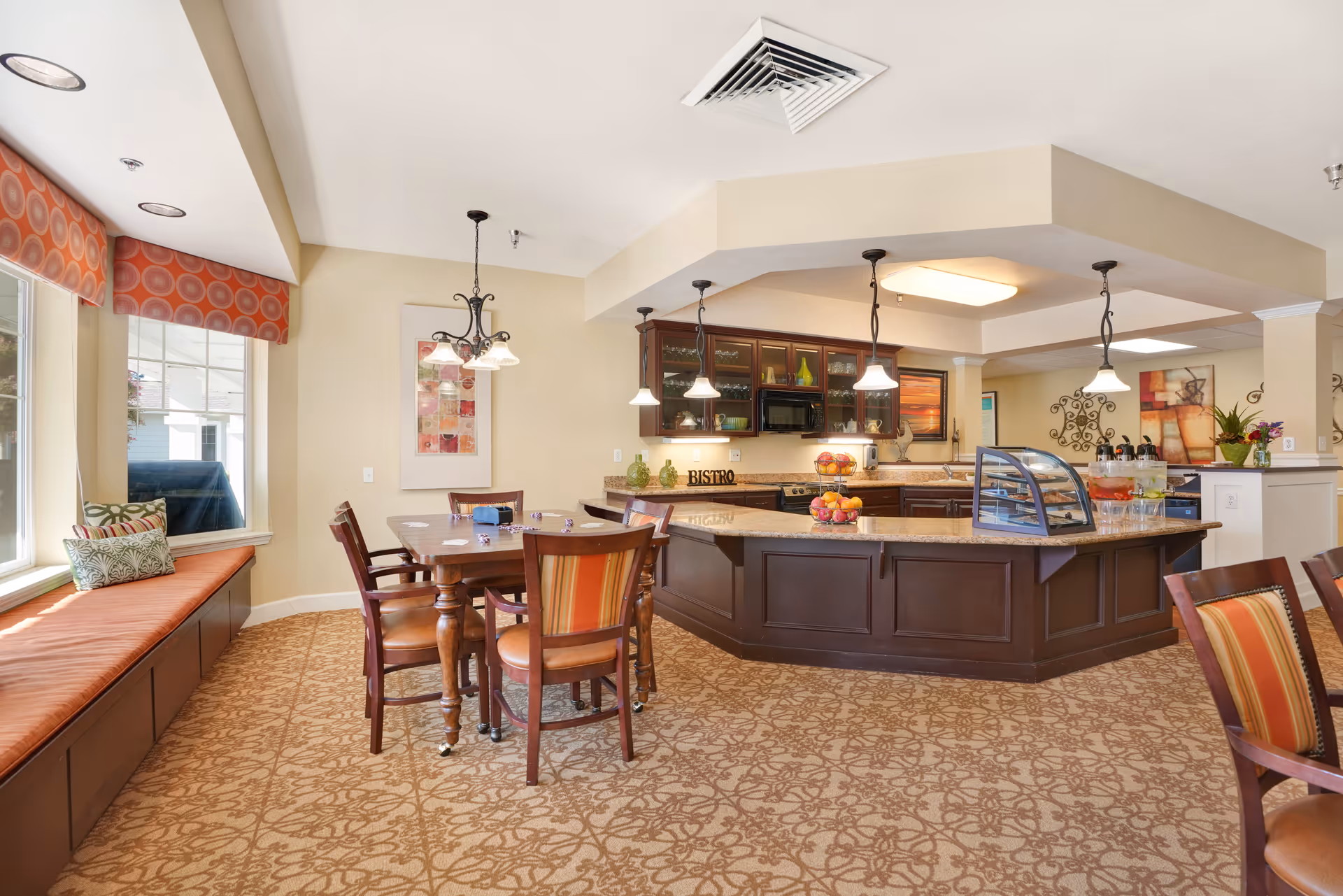 A bright and inviting dining area with a wooden table and chairs, a cushioned window bench with patterned pillows, and a kitchen island with pendant lights hanging above. The kitchen area features dark wood cabinetry, a microwave, and decorative items including a bowl of fruit and a sign that says 'BISTRO'. The room has beige walls and patterned carpet flooring.