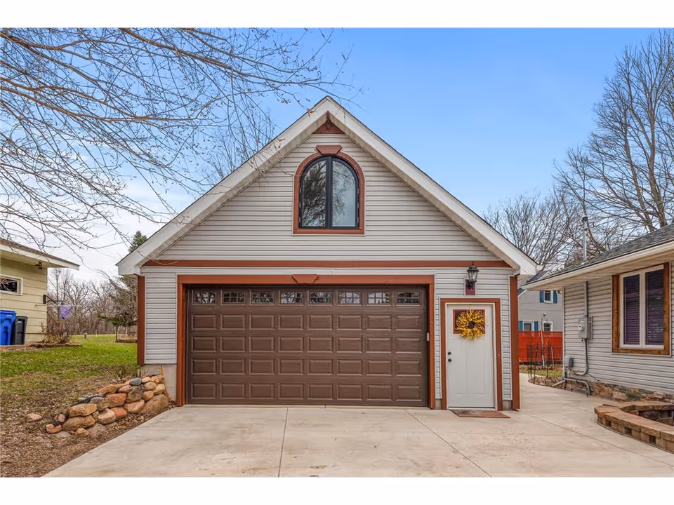 Front view of a detached garage with a brown sectional garage door, adjacent white entry door decorated with a wreath, and a concrete driveway.