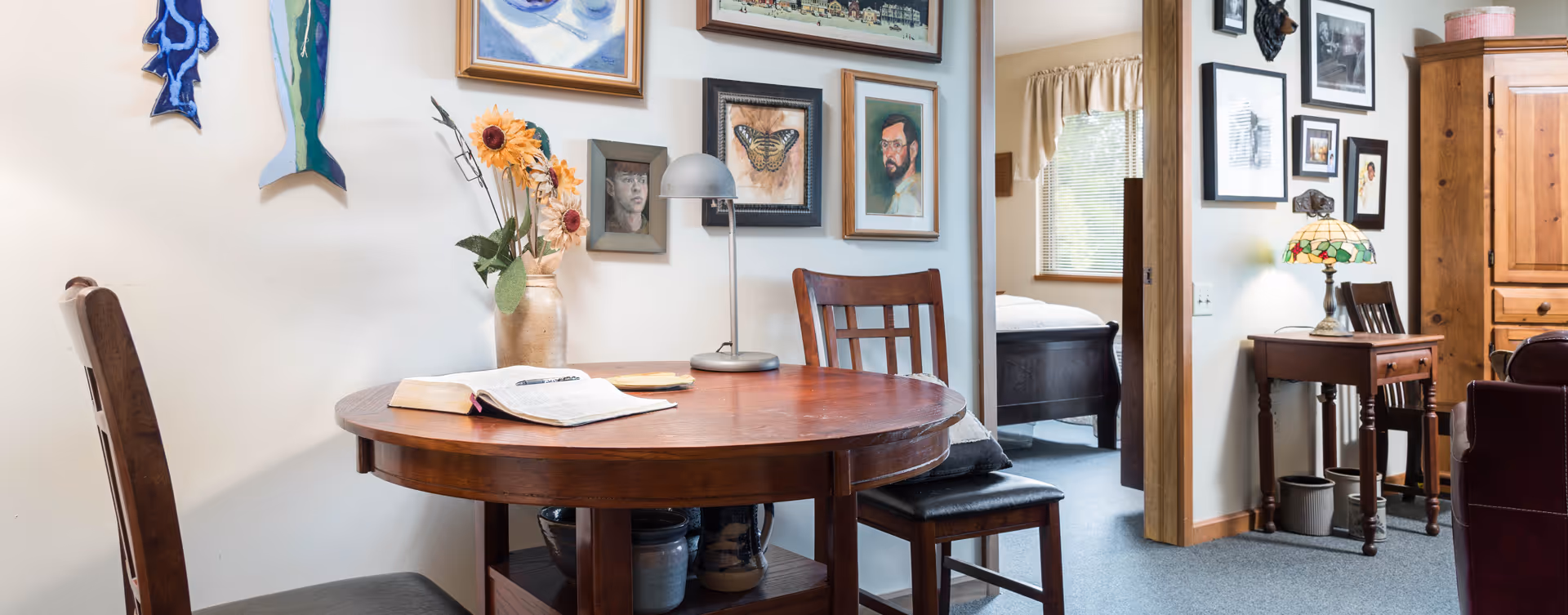 Small dining nook with a round wooden table and chairs, wall art, and a view into a bedroom and adjoining living area.