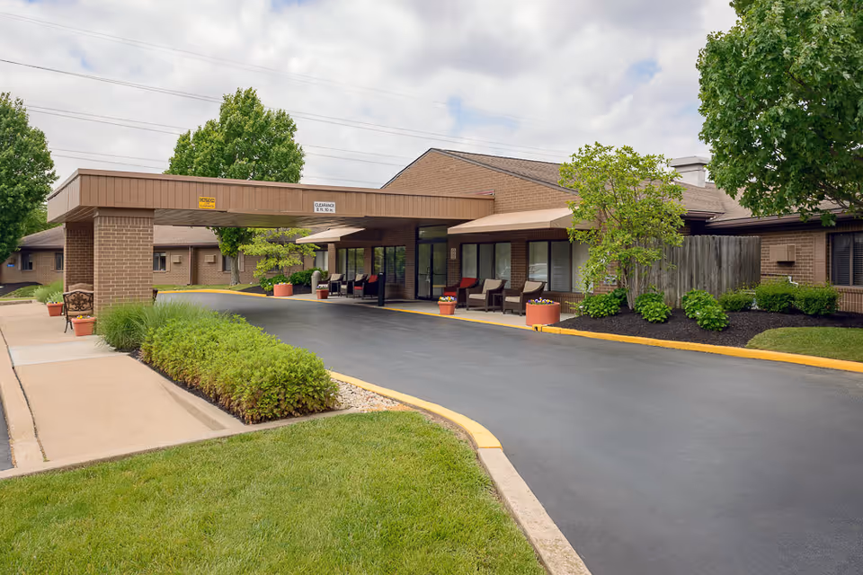 Exterior view of Mayfair Village Nursing Care Center showing the entrance driveway with a covered drop-off area, surrounded by well-maintained landscaping including green grass, bushes, and trees under a partly cloudy sky.