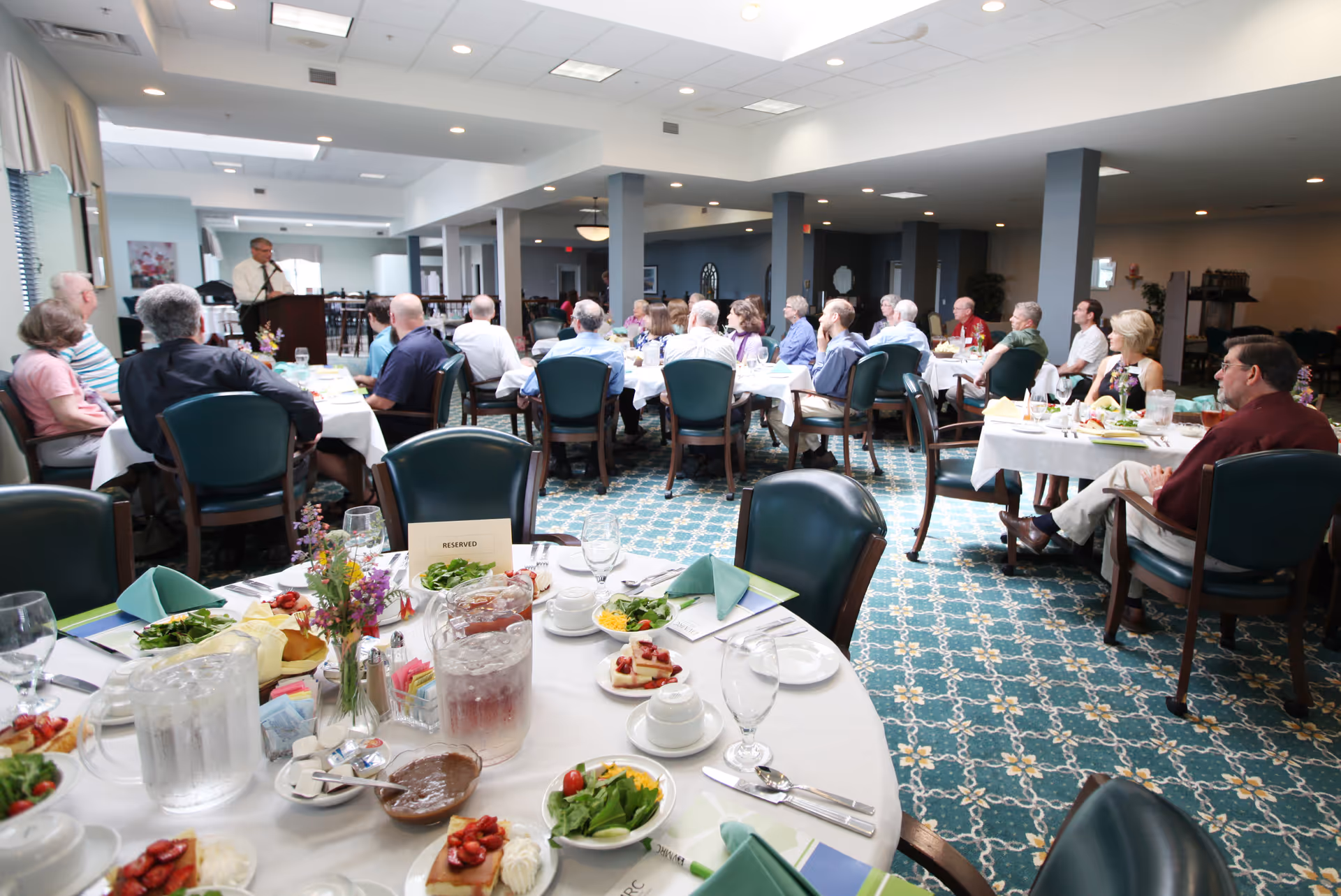 A group of elderly people seated at round tables in a dining room, listening to a man speaking at a podium. The tables are set with plates of salad, desserts, pitchers of water, and floral centerpieces. The room has a patterned carpet and bright overhead lighting.