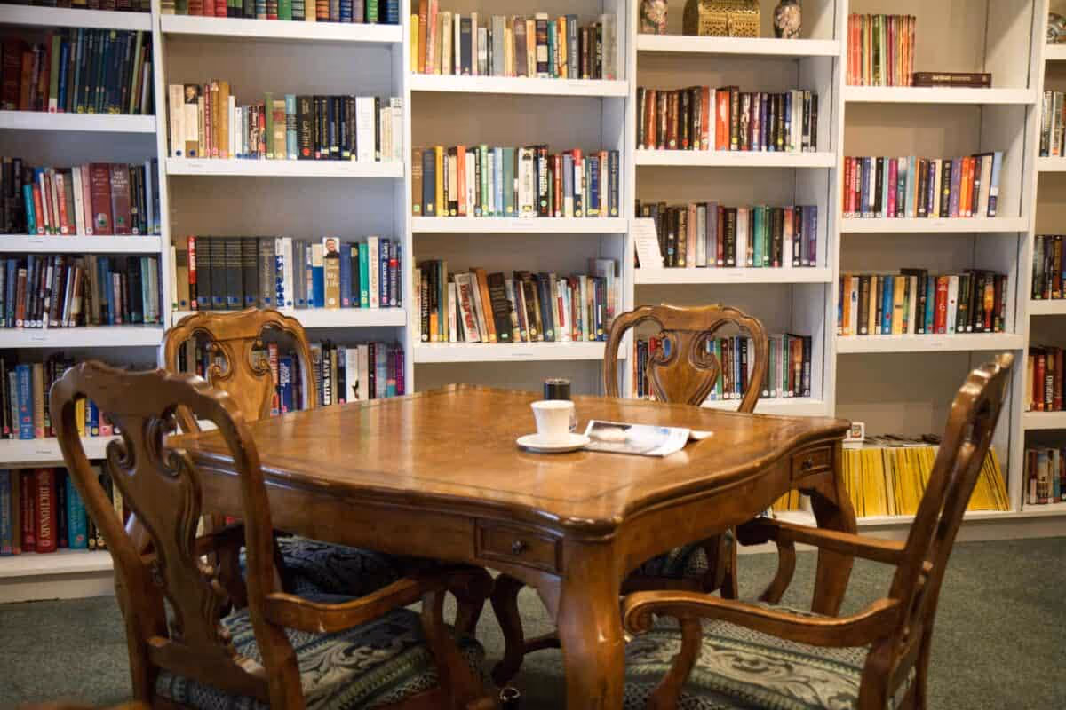 A cozy reading area with a wooden table surrounded by four ornate wooden chairs. Behind the table are white bookshelves filled with a variety of books. On the table, there is a white cup and saucer along with a folded newspaper.