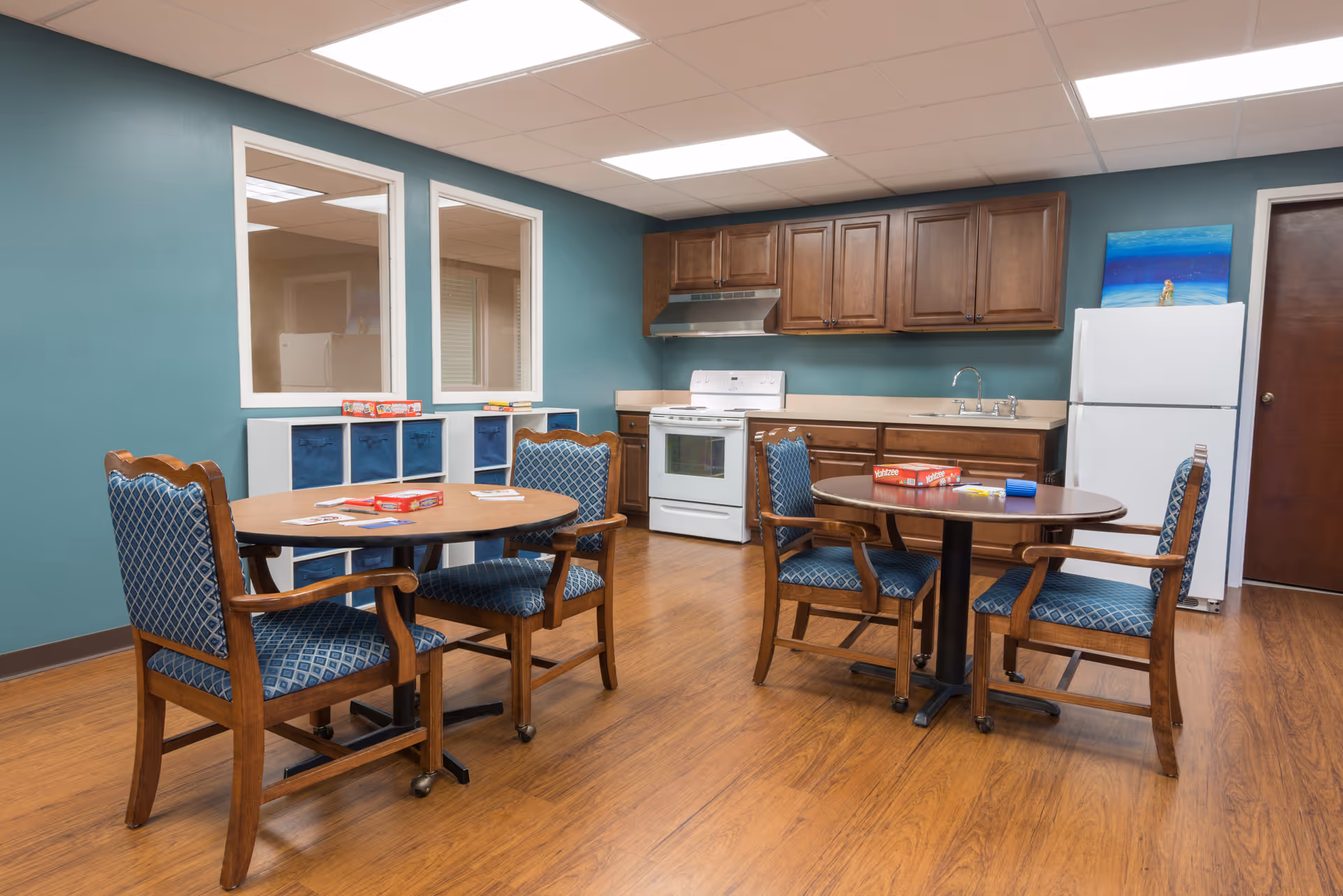 A cozy kitchen and dining area with wooden cabinets, a white stove, a white refrigerator, and two round tables each surrounded by four wooden chairs with blue patterned cushions. The walls are painted teal, and there are two windows on the left wall. Various board games are placed on the tables and a white storage unit against the wall.