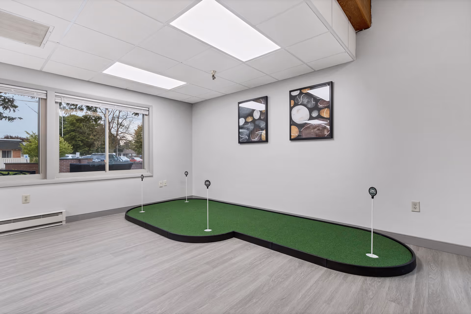 Indoor room with a small putting green for golf practice, featuring four holes with flags. The room has light gray walls, a large window showing an outdoor view with trees and parked cars, two abstract art paintings on the wall, and light wood flooring.