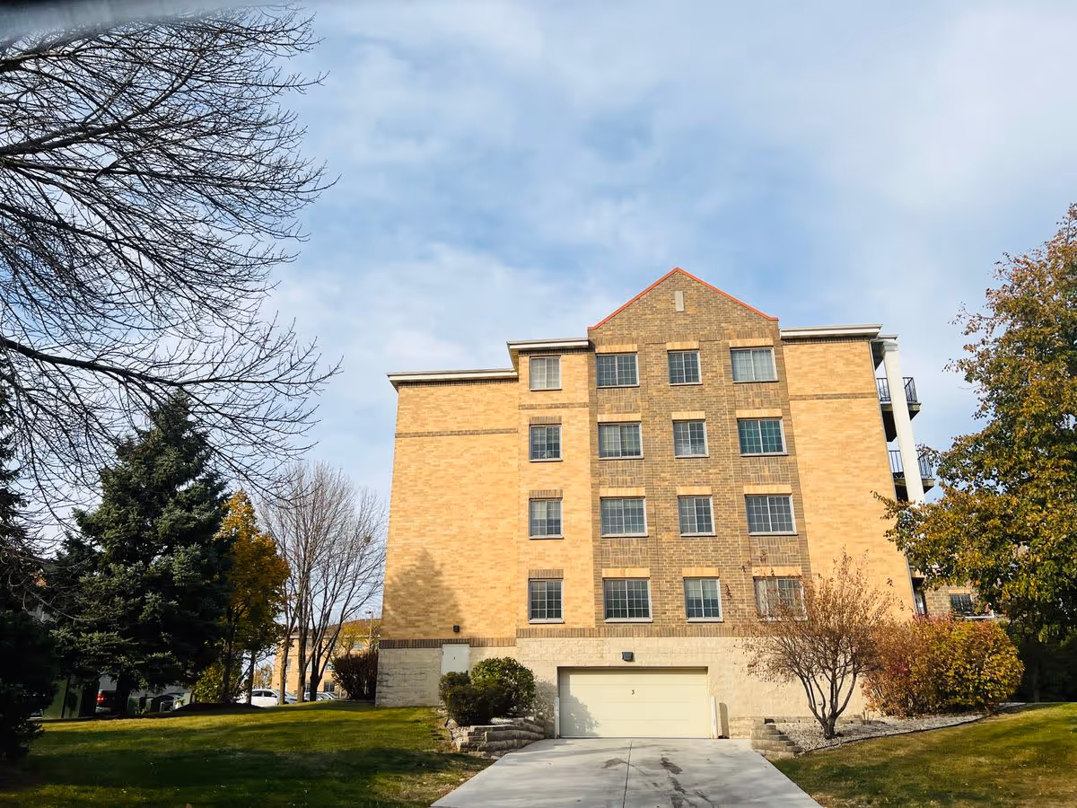 Multi-story brick residential building with a garage and driveway, surrounded by lawn and trees under a partly cloudy sky.