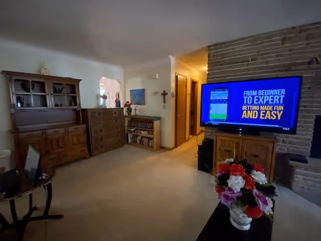 A cozy living room in Richfield Senior Suites featuring wooden furniture including a hutch, dresser, and bookshelf. A flat-screen TV is mounted on a stone wall displaying a blue screen with text. A vase with colorful artificial flowers is on a table in the foreground. The room has beige carpeting and light-colored walls with some decorations including a cross and framed pictures.