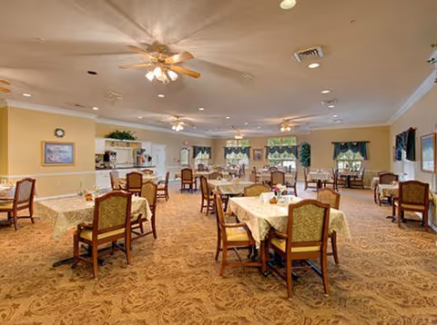 Spacious dining room with multiple tables covered in tablecloths, upholstered chairs, ceiling fans, and warm lighting.