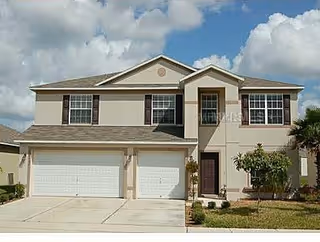 Two-story beige house with a three-car garage, multiple windows, a front door with a small porch, and a small lawn with a tree and bushes under a partly cloudy sky.