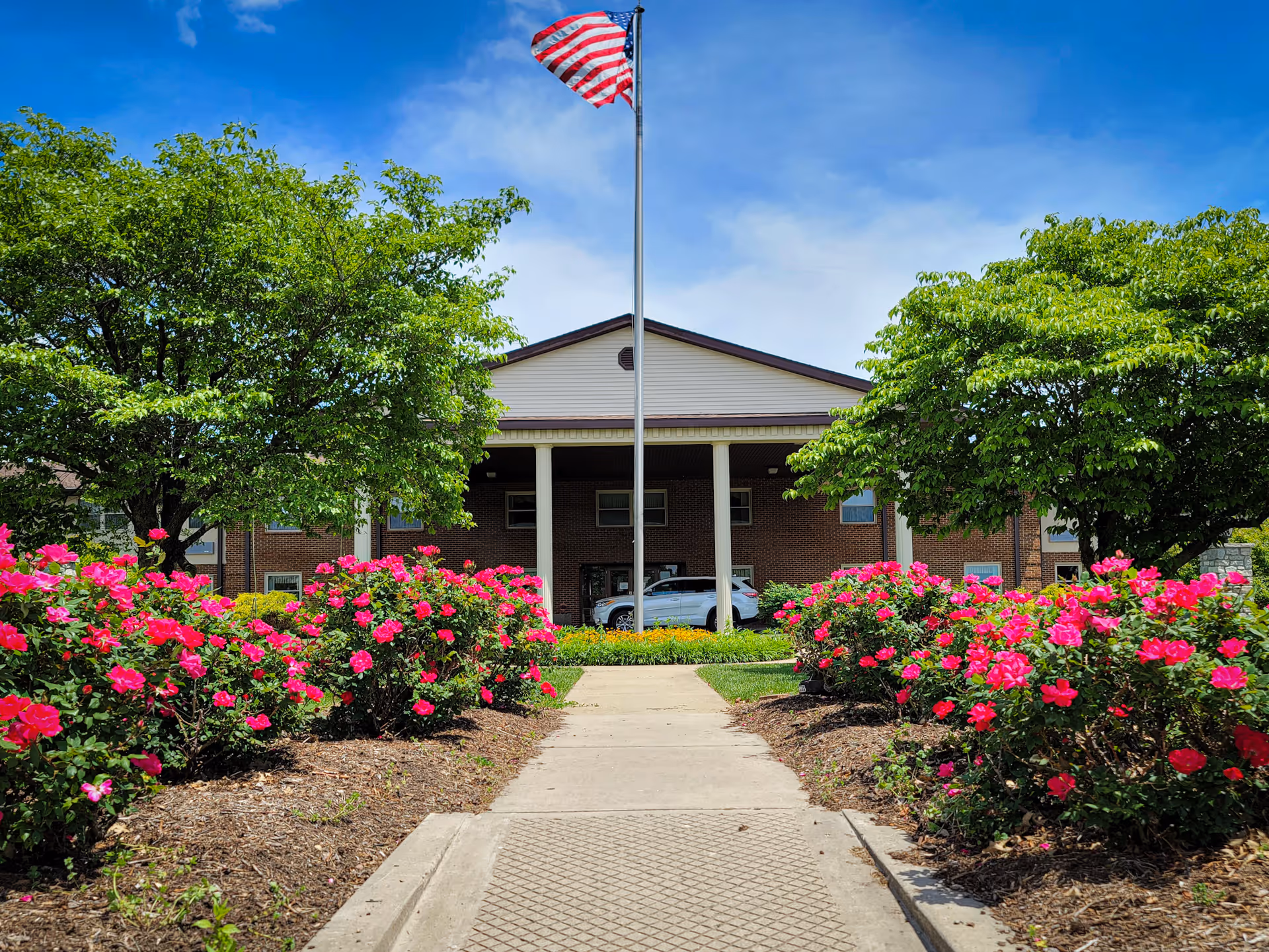 Pathway leading to the entrance of a brick building with white columns, flanked by green trees and vibrant pink flowers, with an American flag flying on a flagpole in front under a blue sky.