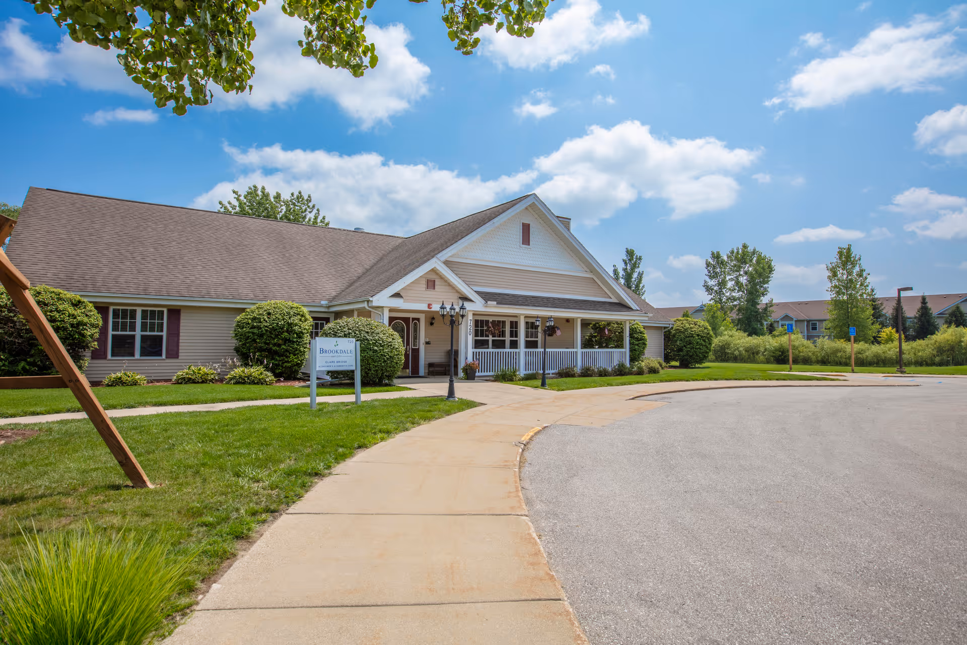 Exterior view of a single-story building with a gabled roof, beige siding, and a white porch railing. There is a paved walkway leading to the entrance, surrounded by green grass and bushes. The sky is blue with scattered clouds.