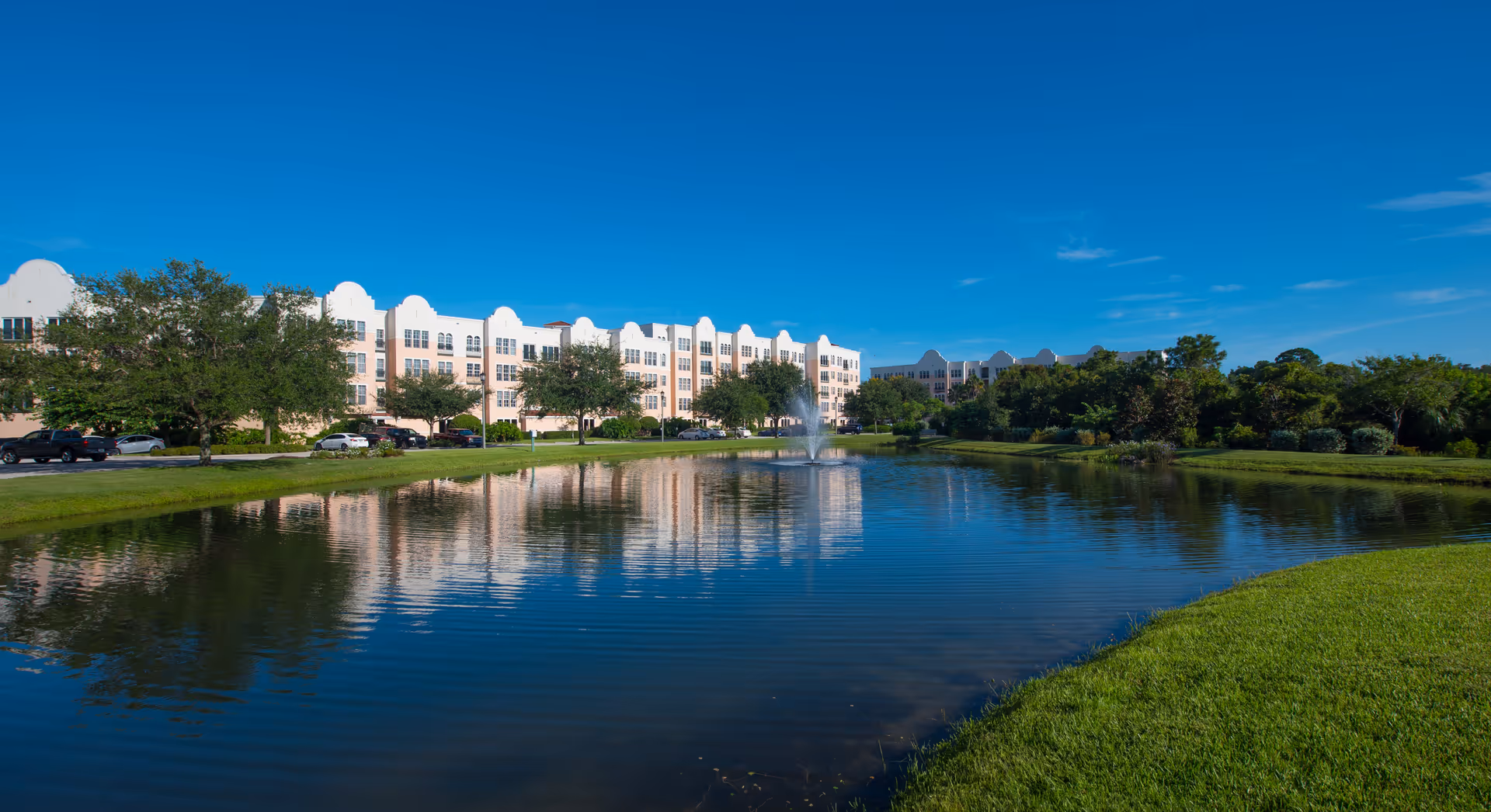 A large pond with a water fountain in the center, surrounded by green grass and trees. In the background, there is a multi-story building with a light-colored facade under a clear blue sky.