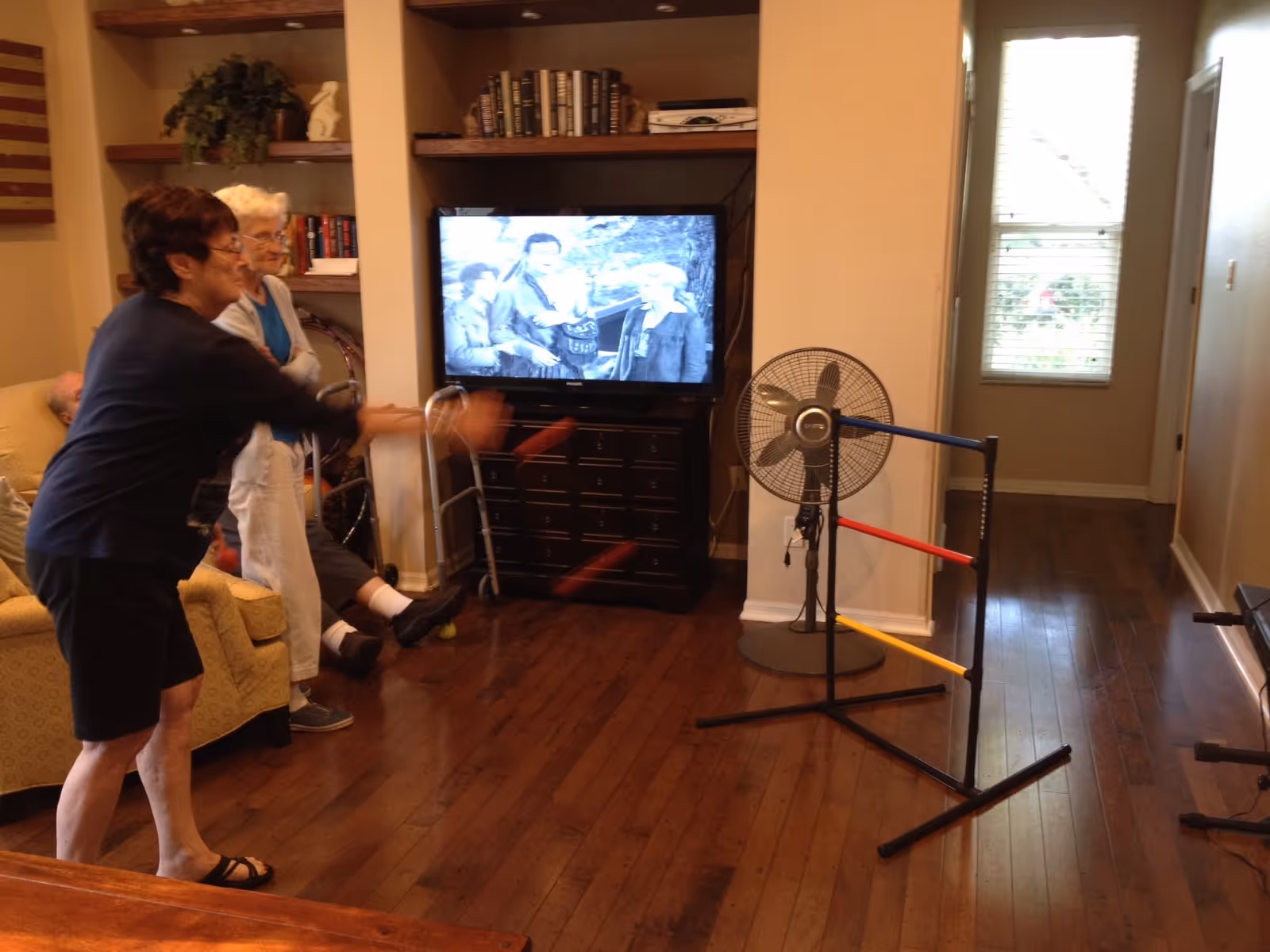 Two elderly women playing a ring toss game in a living room with wooden floors. One woman is actively tossing rings while the other watches. There is a television on a stand showing a black and white program, a standing fan, and a walker nearby. The room has shelves with books and decorations, and a window letting in natural light.
