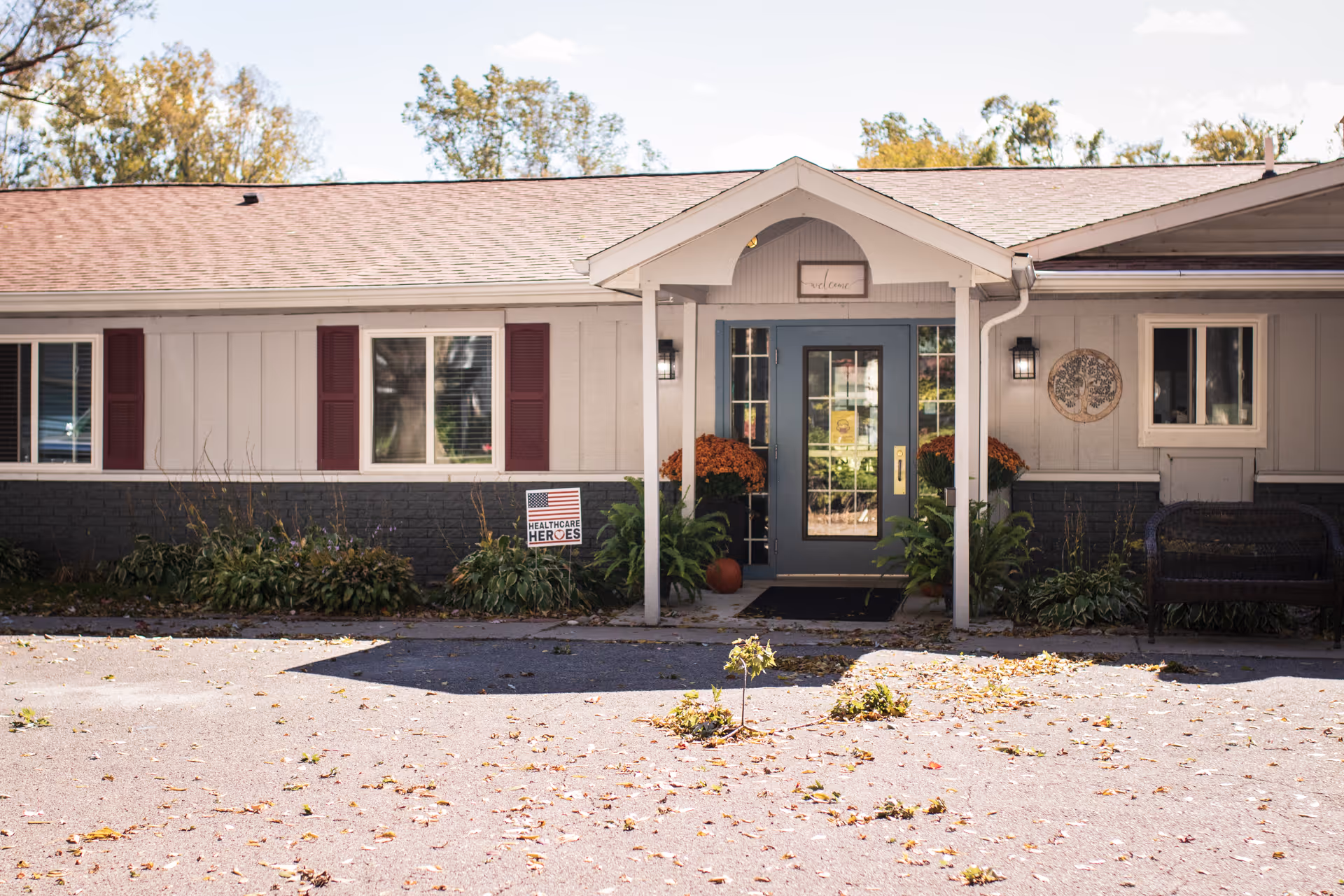 Front entrance of a single-story senior living facility with a covered porch, blue door, potted plants, and a sign that reads 'Healthcare Heroes' in the garden bed. The building has beige siding with maroon shutters and a small bench to the right of the door.