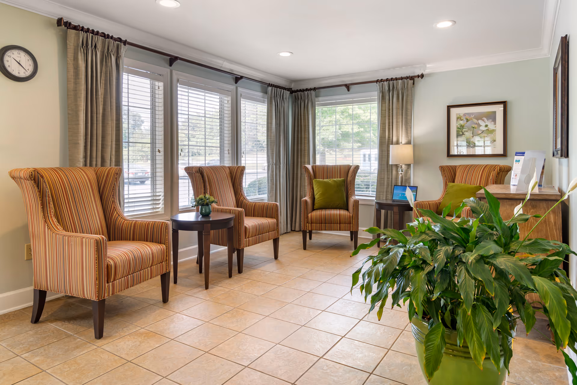 A bright sitting area with four striped armchairs arranged around two small round wooden tables. The room has large windows with blinds and beige curtains, a wall clock, a framed floral picture, a floor lamp, and a large green potted plant in the foreground. The floor is tiled with light-colored tiles.