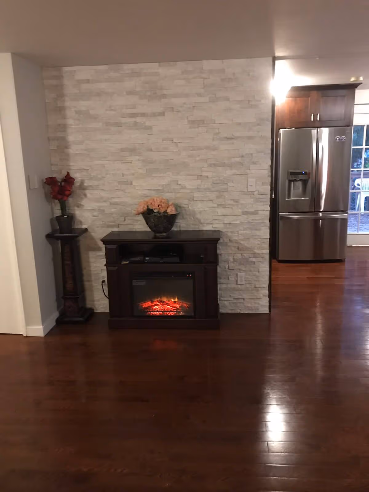 Interior living area with an electric fireplace against a stone accent wall, hardwood floors, and a stainless refrigerator visible to the right.