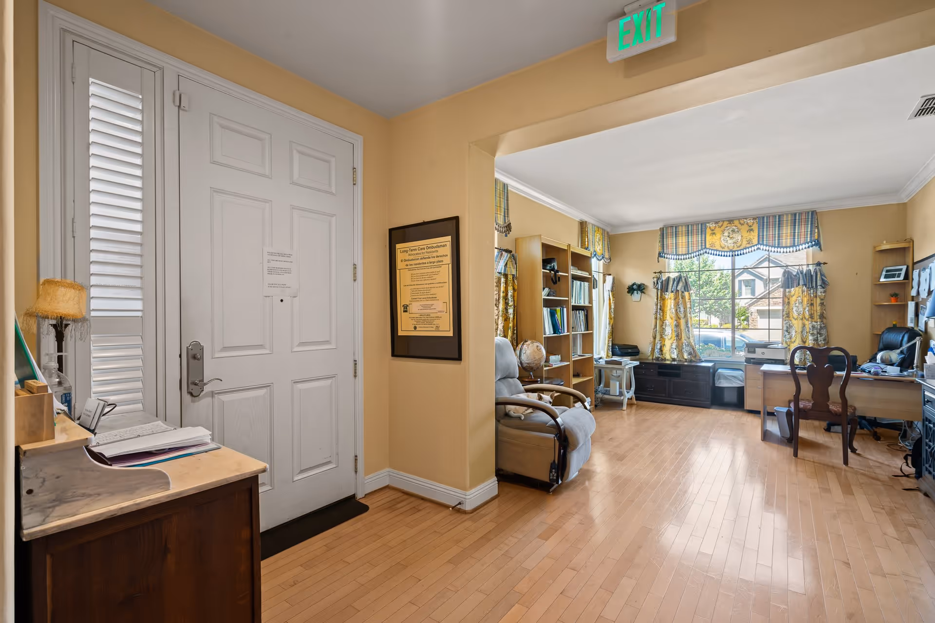 Interior view of a room in Aunt Dottie's Place showing a white front door on the left, a small wooden table with papers and a lamp, and an adjacent office area with bookshelves, a reclining chair, a desk with a computer, and large windows with yellow and blue patterned curtains letting in natural light.