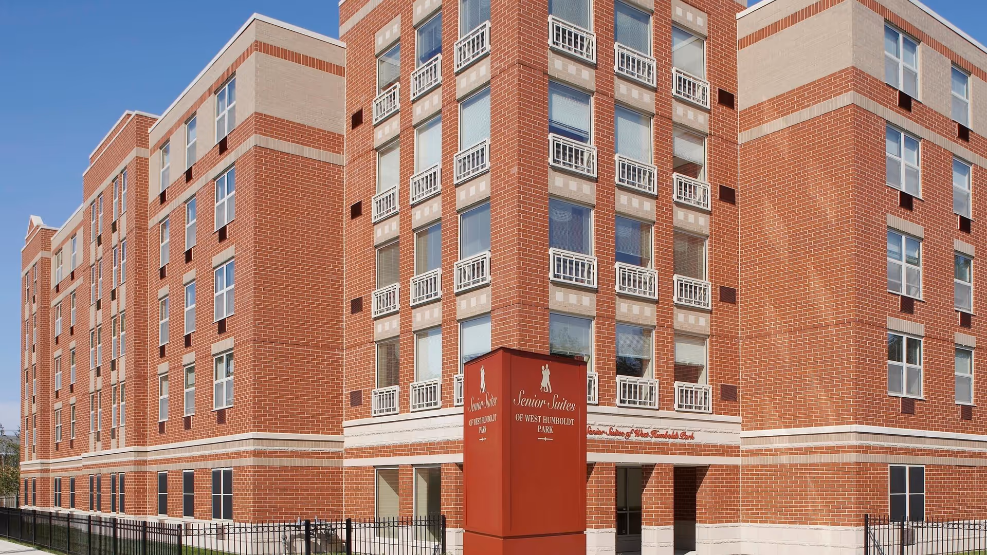 Front exterior of a multi-story red-brick senior living building with a sign reading 'Senior Suites of West Humboldt Park' and a fenced entrance.
