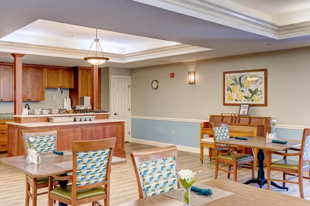 A dining area in a senior living facility featuring wooden tables and chairs with patterned cushions. The room has light-colored walls with a blue wainscoting, a framed lemon-themed artwork, and a small piano against the wall. In the background, there is a kitchen area with wooden cabinets and a counter with various items on it. The ceiling has recessed lighting and a hanging light fixture.