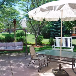 Patio area with an umbrella-covered table, chairs, a swing and a white bench set in a landscaped garden.