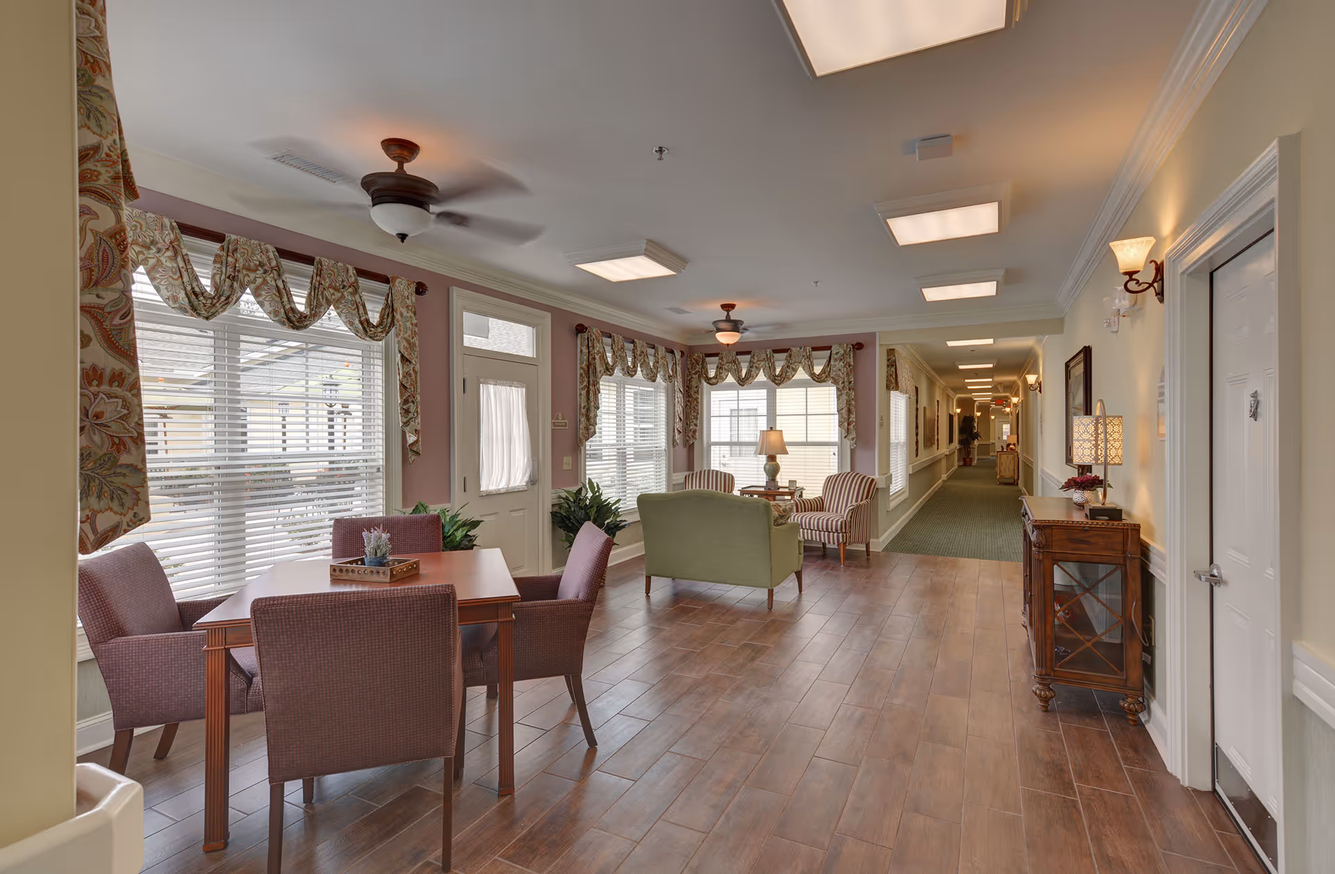 A bright and spacious common area in a senior living facility with wooden flooring, a table with four chairs, two striped armchairs, a green loveseat, and large windows with patterned valances. Ceiling fans and lamps provide lighting, and a long hallway extends in the background.