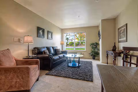 Living room with a sofa, armchair, coffee table on a rug and a large window overlooking greenery.