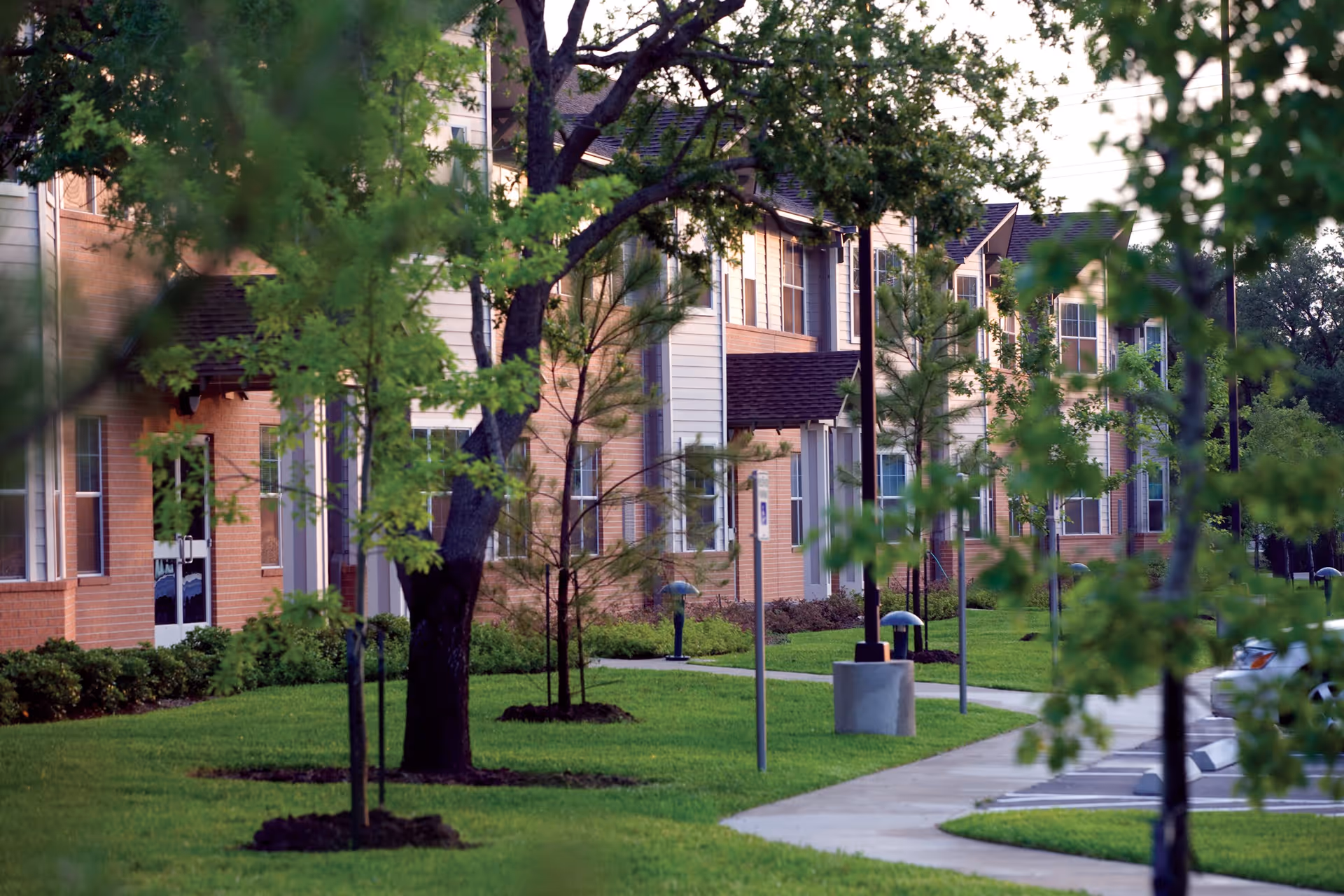 View of the exterior of a senior living facility named Holly Hall, showing a row of two-story buildings with brick and siding facades, surrounded by green grass, young trees, and a paved walkway.