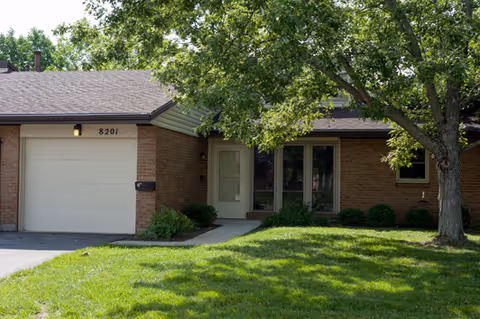 Front exterior of a single-story brick residence with an attached garage, entry door, windows, and a tree-shaded lawn.